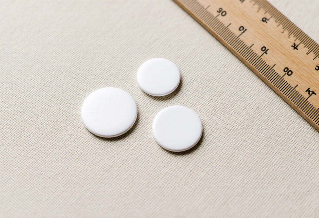 Two sizes of plain round pin badges on linen beside a wooden ruler