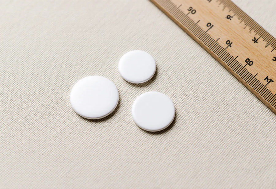 Two sizes of plain round pin badges on linen beside a wooden ruler