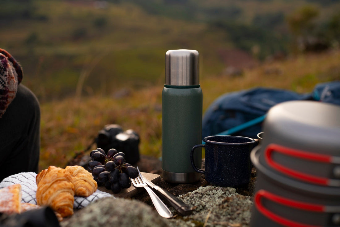 A sage-green vacuum flask and enamel mug at a hillside picnic — the kind of setup that earns its name only with a pre-warmed bottle