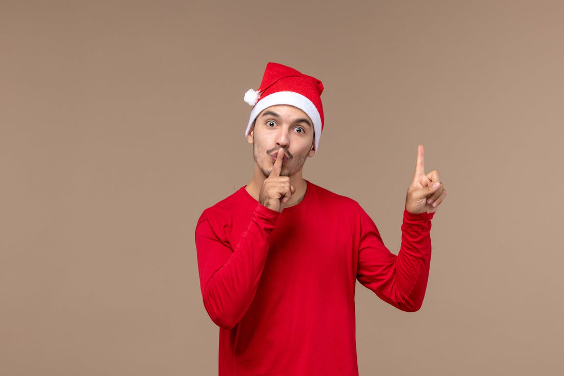 A young man in a Santa hat with a finger to his lips — the universal 'keep it quiet' gesture, the visual setup for an office Secret Santa