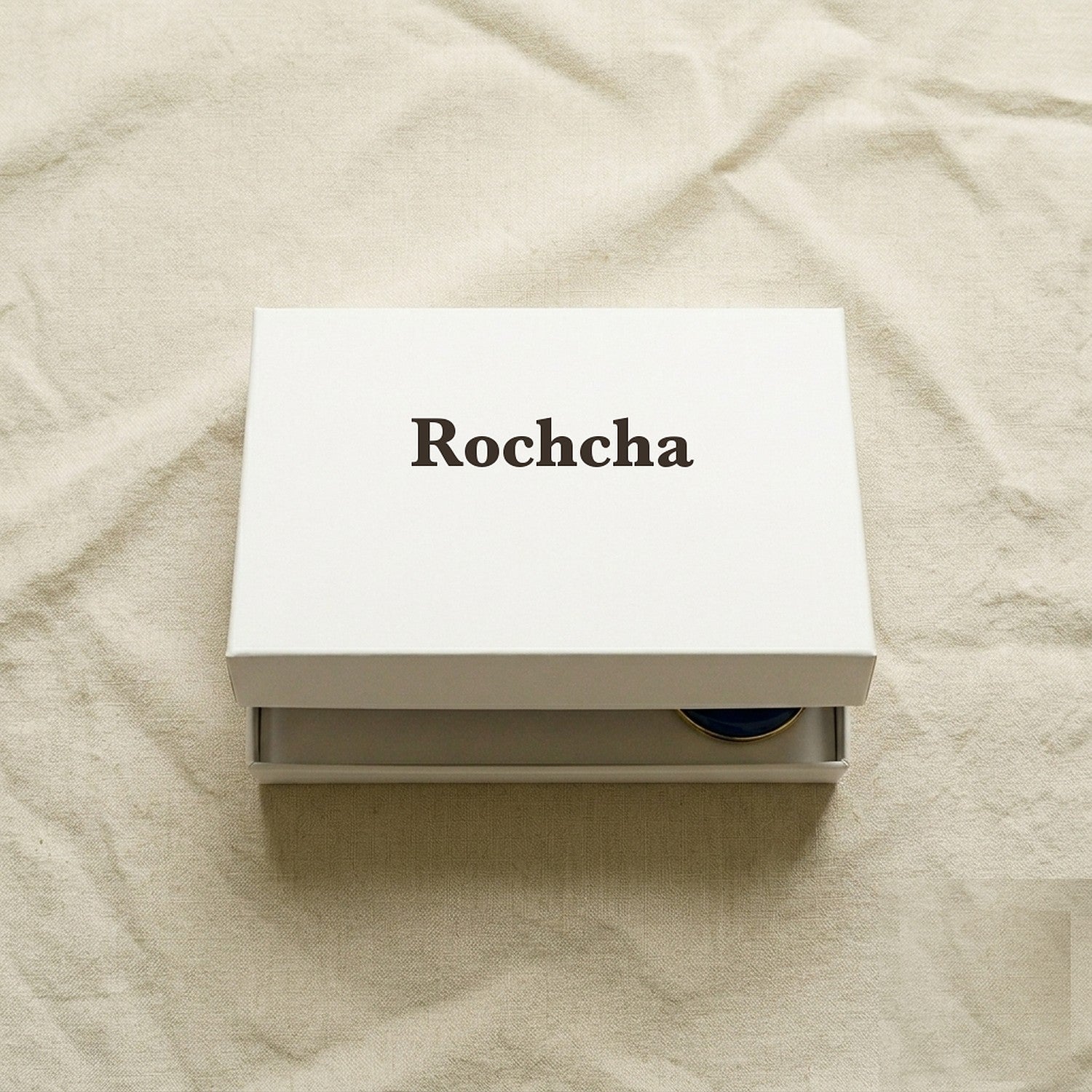 Rochcha-branded white flat product box on cream linen, slightly open to reveal the evil-eye charm set inside.