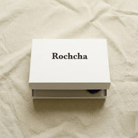Rochcha-branded white flat product box on cream linen, slightly open to reveal the evil-eye charm set inside.