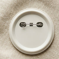Back of a Mawlaii Rochcha Custom Print Badge — matte off-white plastic shell with stainless steel pinback hardware, resting on warm cream linen.