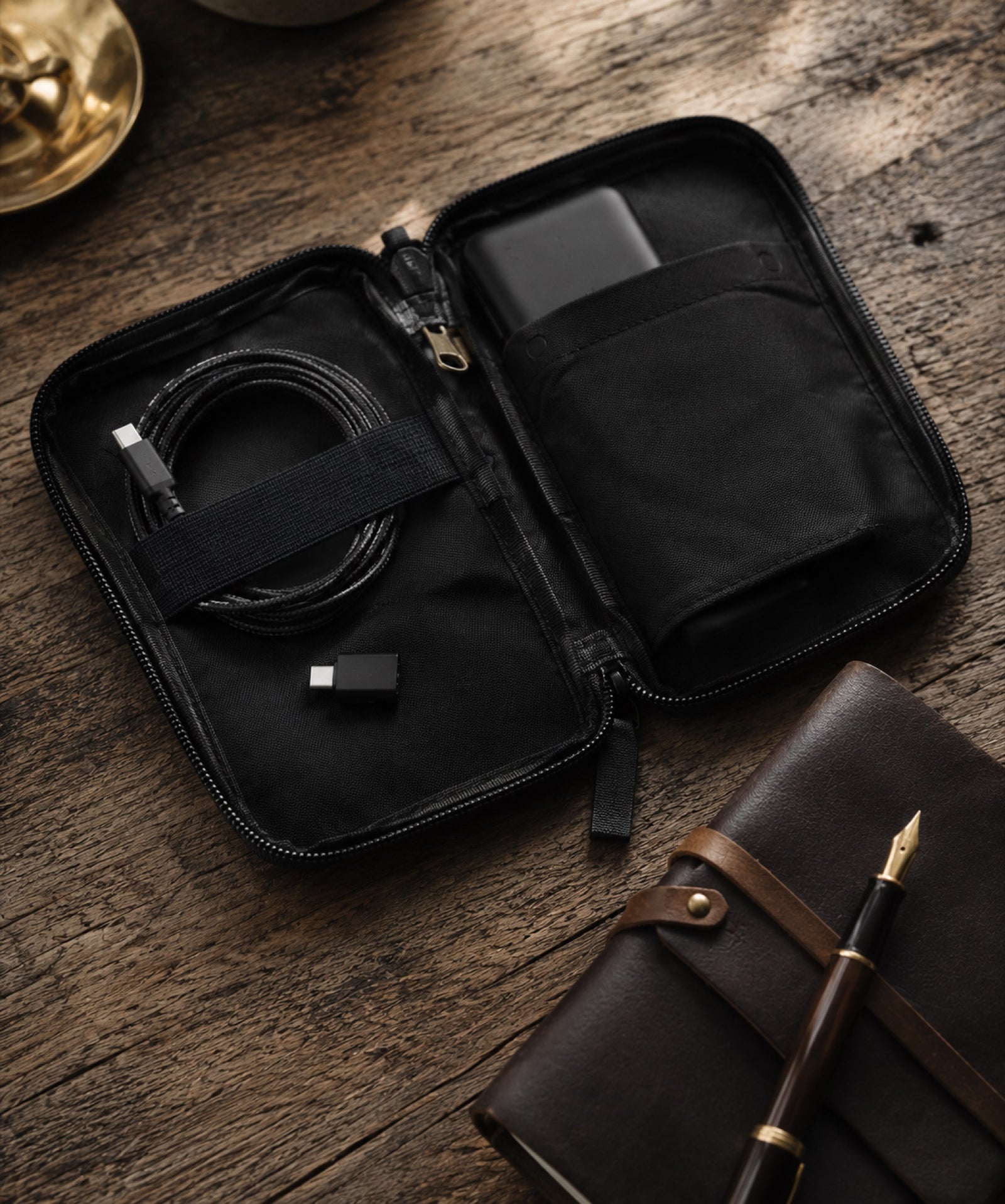 Kaiyo Tokyo — an open tech pouch on a weathered wooden table with coiled cable, dongle, power bank, beside a leather notebook and fountain pen